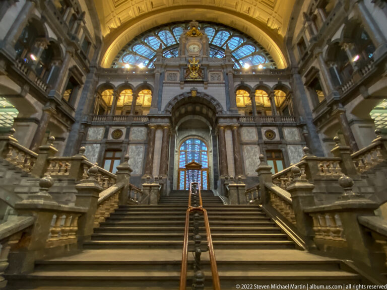 Frontal view of Antwerpen-Centraal’s broad twin staircase beneath a large arched fanlight and ornate stone architecture.
