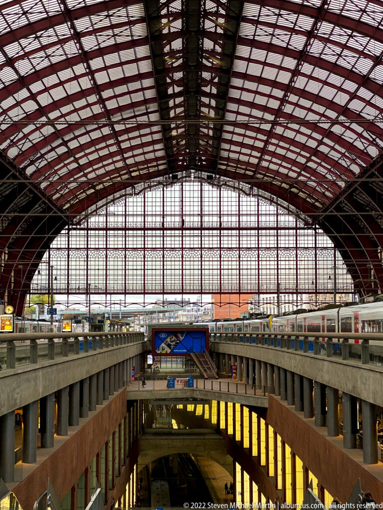 Wide interior of Antwerpen-Centraal with arched steel-and-glass train shed above platforms and lower concourse levels.