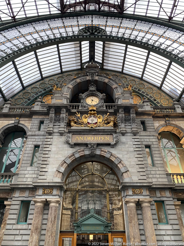 Ornate stone entrance façade of Antwerpen-Centraal beneath a glass-and-iron canopy, with gilded decoration and ANTWERPEN inscription.