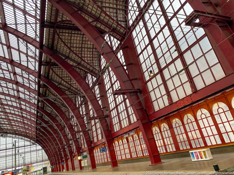 Upward view inside Antwerpen-Centraal showing red iron arches, roof trusses, and tall gridded glass walls.
