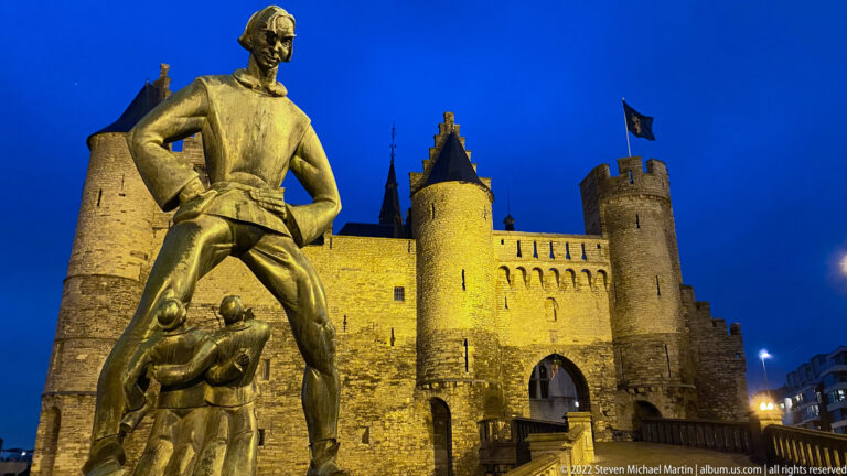 Bronze Lange Wapper statue in front of the illuminated medieval gatehouse and round towers of Het Steen at dusk.