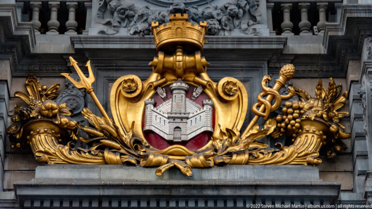 Close-up of gilded Antwerp civic crest surrounded by a trident, caduceus, fruit, leaves, and carved stone ornament.