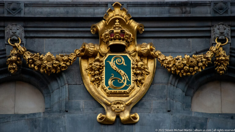 Crowned green-and-gold monogram shield with gilded garlands mounted on the gray stone façade of Antwerpen-Centraal.
