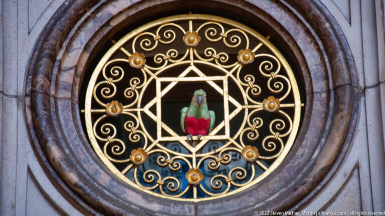 Round gilded grille with scrolling metalwork, geometric star pattern, and a small colorful parrot figure at the center.