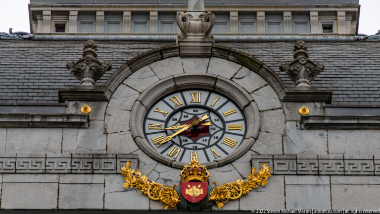 Large Roman-numeral clock set in pale stone beneath a carved crest and above a small shield with gold ornament.