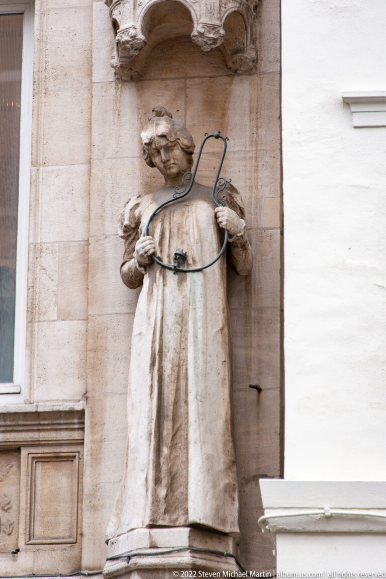 Weathered stone statue of a robed figure in a wall niche, with softened features and a dark cable crossing the torso.