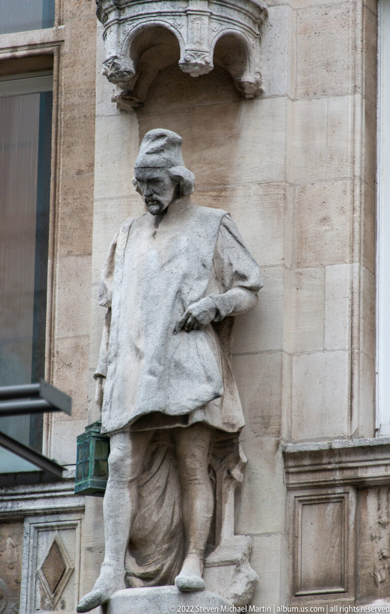 Weathered stone statue of a standing man in broad hat and layered clothing, set within an exterior architectural niche.