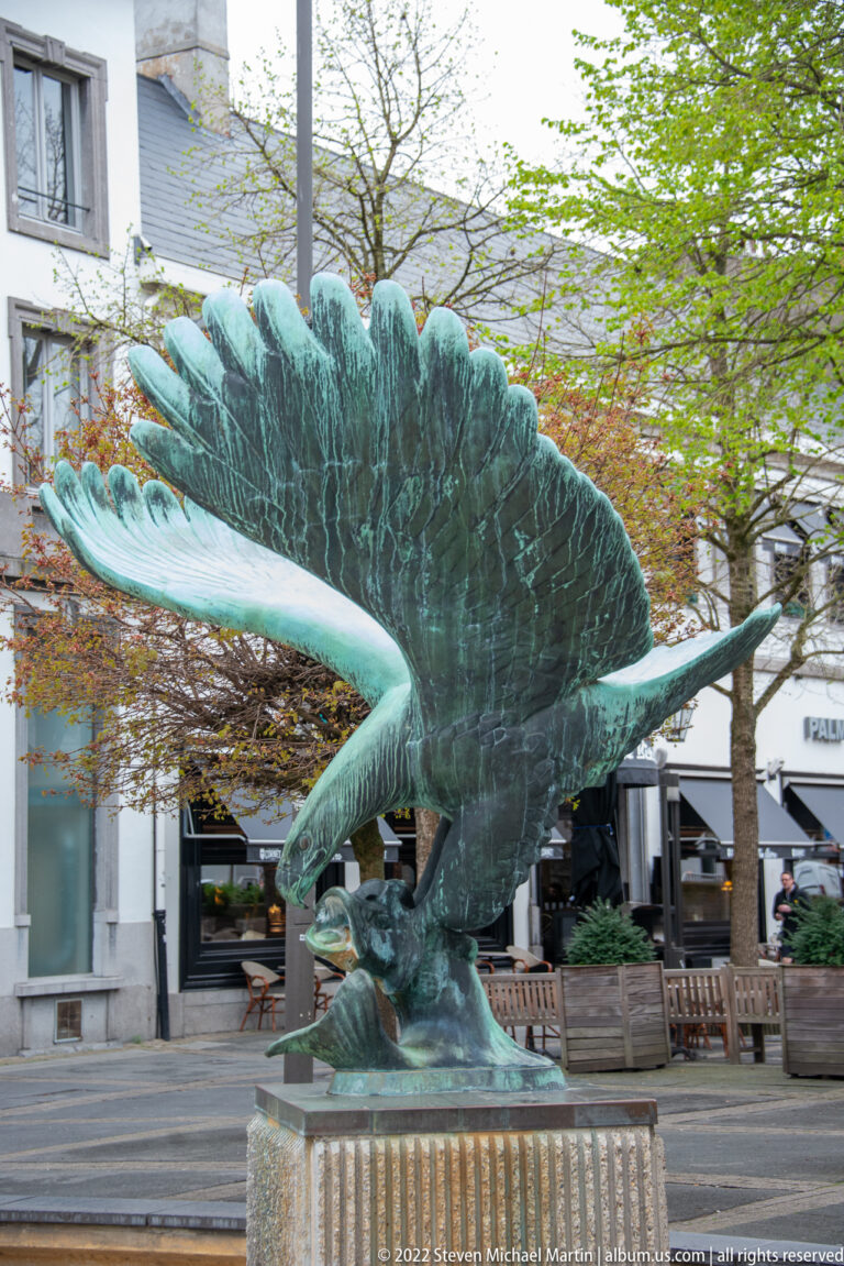 Large green-patinated bronze eagle with wings spread wide on a low pedestal along an Antwerp street.