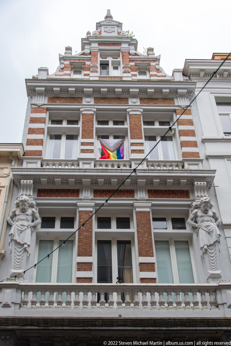 Upward view of ornate brick-and-stone townhouse with sculpted figures, balustrades, stepped gable, and a rainbow flag.