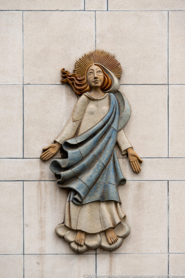 Stylized relief of a robed female figure with blue drapery, outstretched arms, and radiating hair on a pale wall.