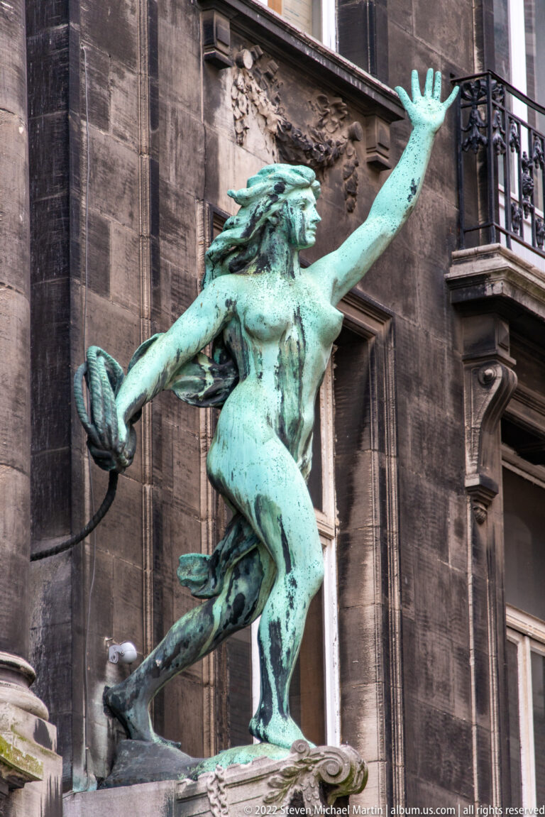 Green-patinated bronze nude male figure in a twisting pose, mounted against a dark stone building façade.