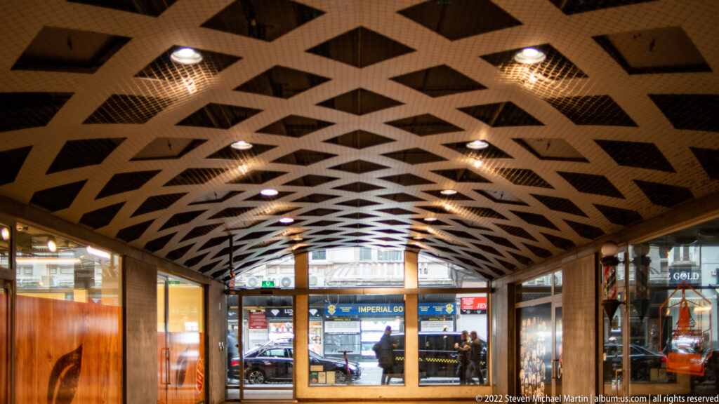 Covered station passage with diamond-patterned ceiling, shopfronts, and glass doors opening toward the street.