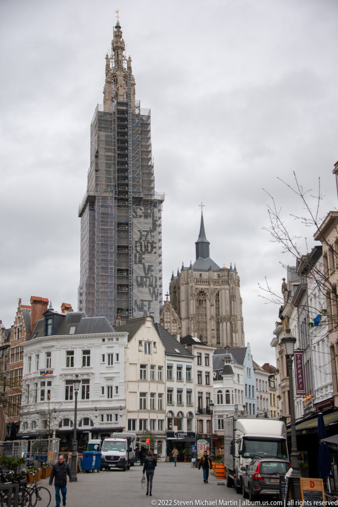 Antwerp street scene with shops and pedestrians leading toward the scaffolded Gothic tower of the Cathedral of Our Lady.