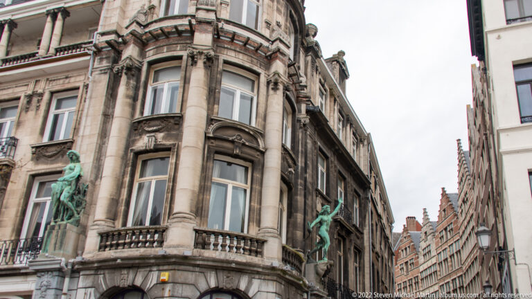 Upward view of ornate rounded stone corner building with bay windows, carved details, balustrades, and green bronze figures.