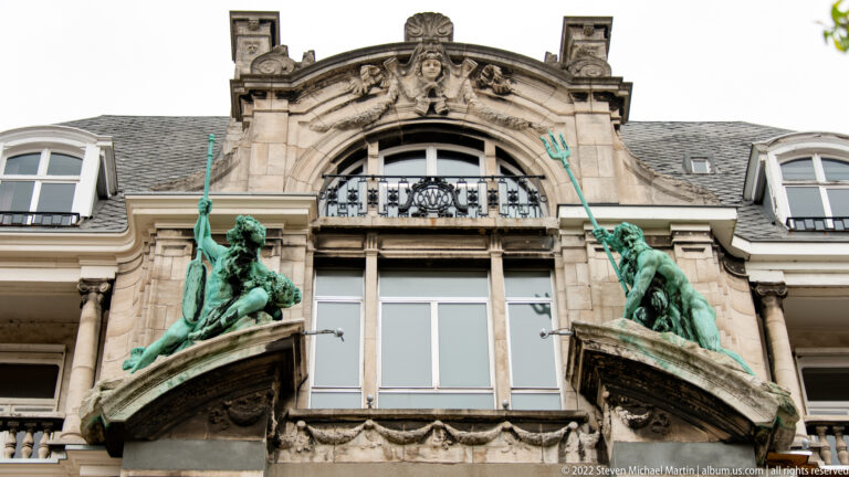 Two green bronze figures holding tridents flank windows and an ornate curved gable on a historic Antwerp façade.