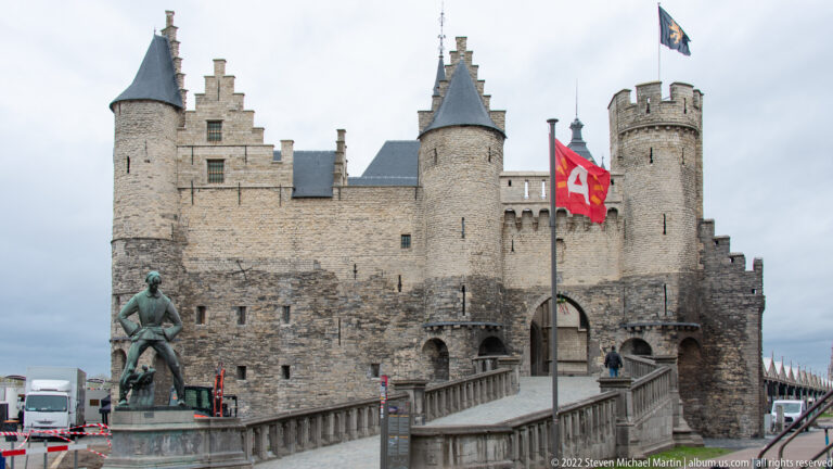 Front approach to Het Steen with round stone towers, central gate, flags, raised bridge, and a bronze statue at left.