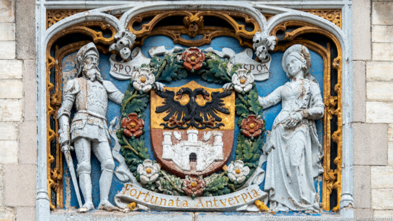 Painted heraldic relief with crowned Antwerp shield, flanking figures, flowers, and the motto Fortunata Antwerpia.