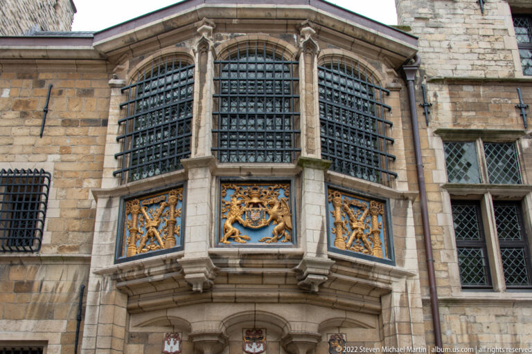Stone oriel window with heavy iron grilles above an entrance, decorated below with three gilded heraldic panels.