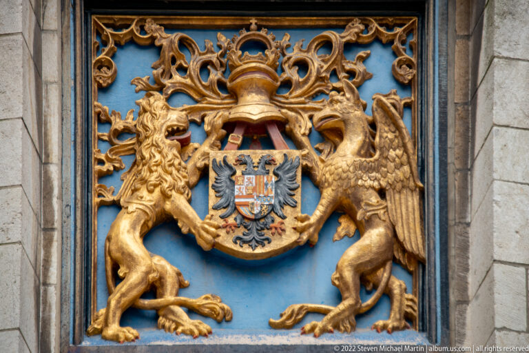 Ornate gilded heraldic panel with lion and winged supporter flanking a crowned shield bearing a black double-headed eagle.