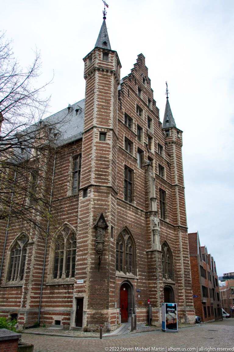 Large historic brick building with stepped gables, corner tower, mullioned windows, and pointed Gothic openings.