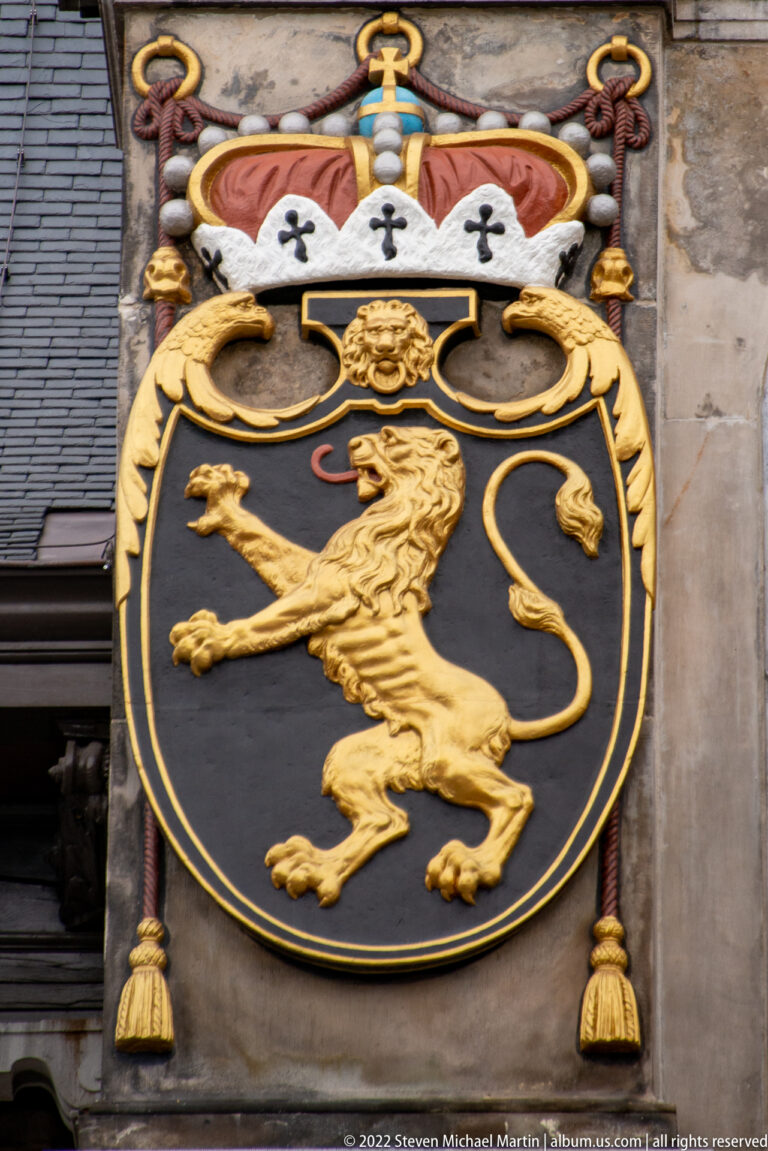 Black heraldic shield with a crowned gold rampant lion, gold mantling, and hanging tassel ornaments on pale stone.