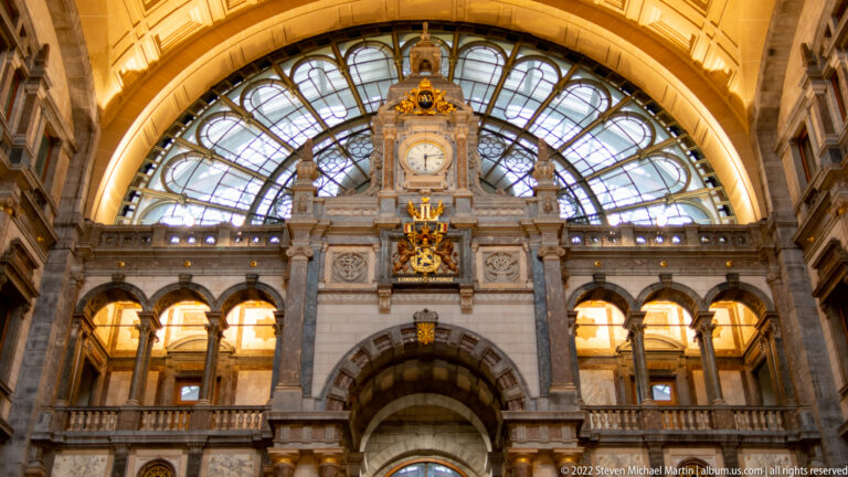 Grand Antwerp-Central Station hall with central clock, gilded ornament, stone arches, and glass roof.