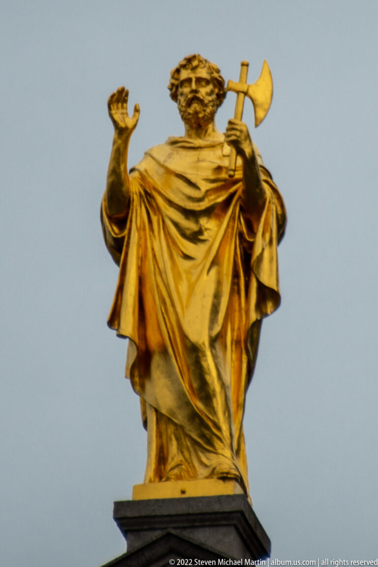 Gilded bearded male statue in draped clothing, one hand raised and a long-handled axe resting at the shoulder.