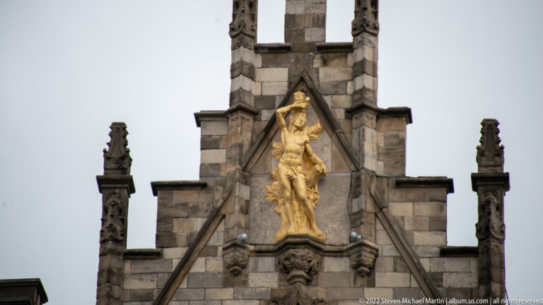 Gilded religious figure standing in a pointed niche on a steep stone gable with Gothic pinnacles.