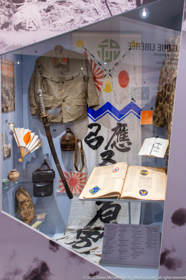 Display case with Japanese wartime uniforms, rising-sun imagery, calligraphic cloths, documents, headgear, and personal equipment.