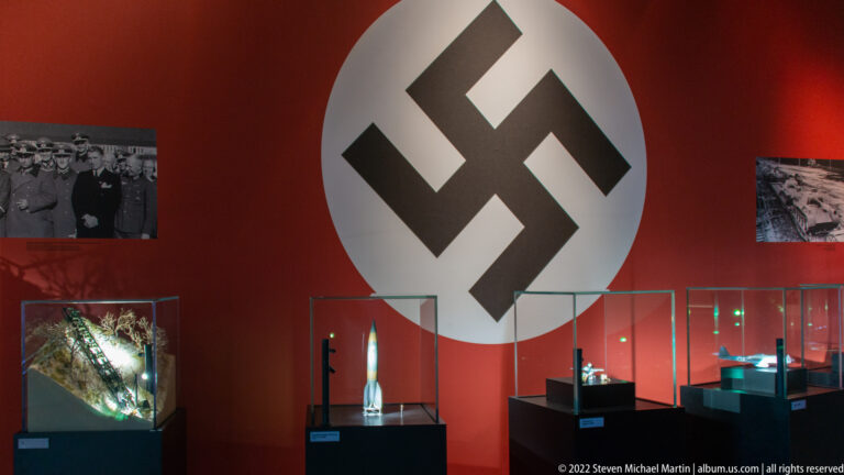 Large black swastika in a white circle on a red museum wall above glass artifact cases and wartime photographs.