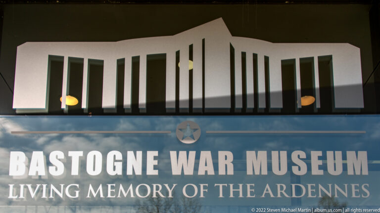 Museum sign reading Bastogne War Museum and Living Memory of the Ardennes below a stylized building silhouette.