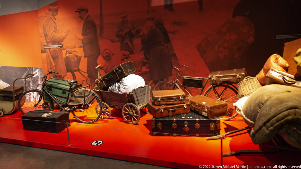 Museum display of bicycles, handcarts, suitcases, trunks, baskets, bedding, and bundled belongings on a red platform.
