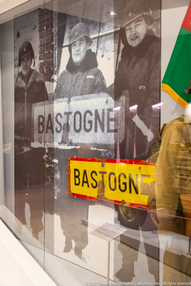 Yellow road sign reading BASTOGNE displayed before a large wartime photograph of soldiers and another Bastogne sign.