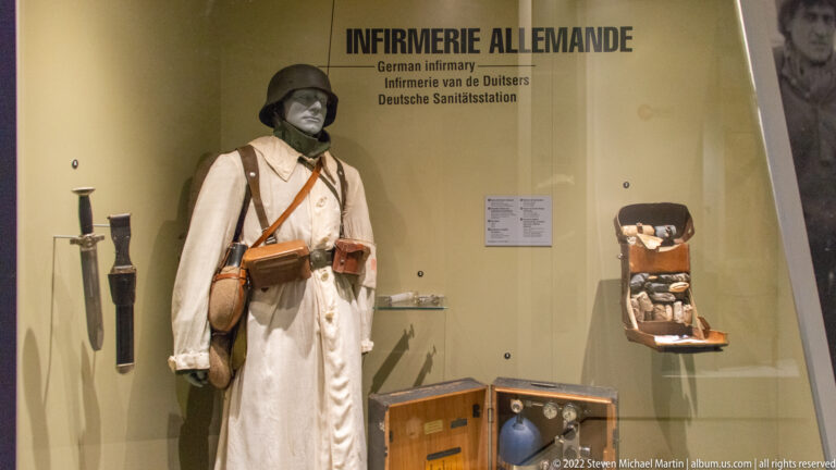 German infirmary exhibit with winter-uniformed mannequin, helmet, pouches, medical tools, containers, and boxed supplies.