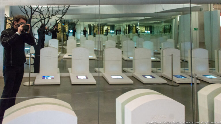 Rows of white grave-marker forms with small screens reflected repeatedly in mirrored walls inside the museum.