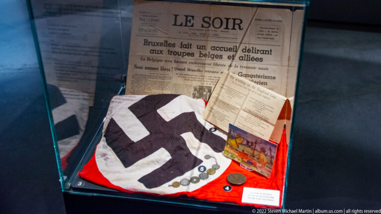 Display case with Le Soir newspaper, printed notices, folded swastika cloth, coins or medals, and colorful liberation-era paper items.