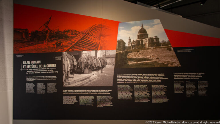 Red-and-black museum panel with wartime photographs, ruined buildings, marching people, and a large domed structure.