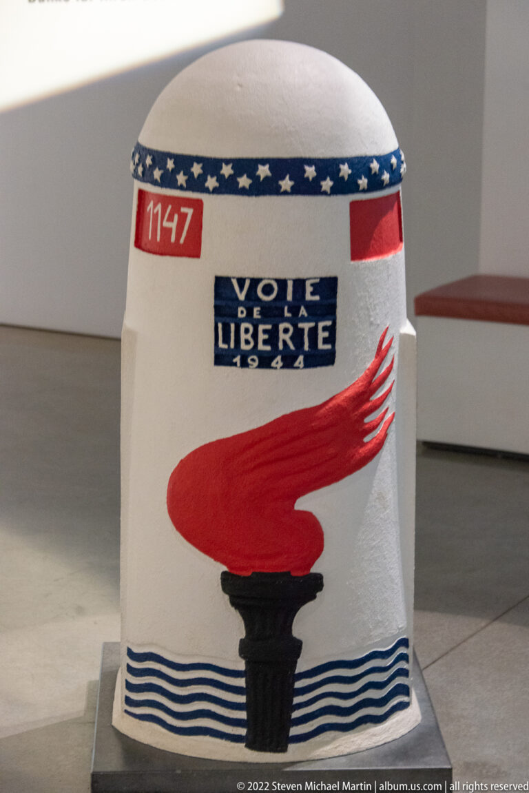 White commemorative marker marked 'VOIE DE LIBERTE' with red torch, blue waves, number 1147, and date 1947.