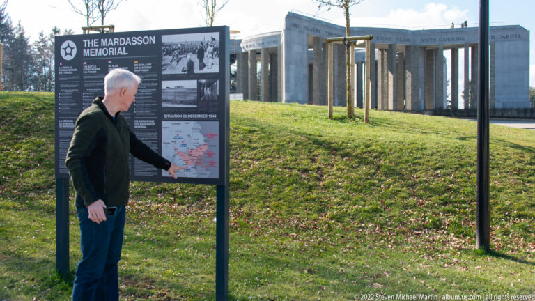 Man reading an outdoor interpretive panel with the Mardasson Memorial visible across the lawn.