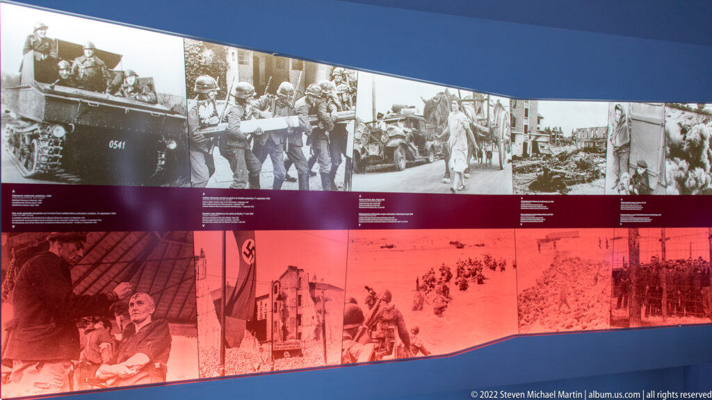 Curved museum wall with rows of black-and-white wartime photographs above a red band of images and captions.