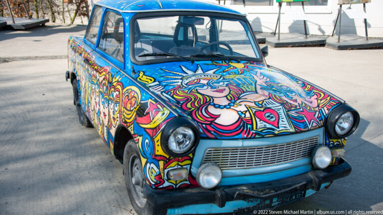 Small blue sedan covered in colorful psychedelic faces, flowers, and graphic patterns.