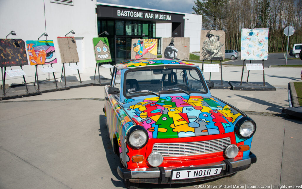 Colorfully painted compact car parked before the Bastogne War Museum entrance and outdoor artworks.