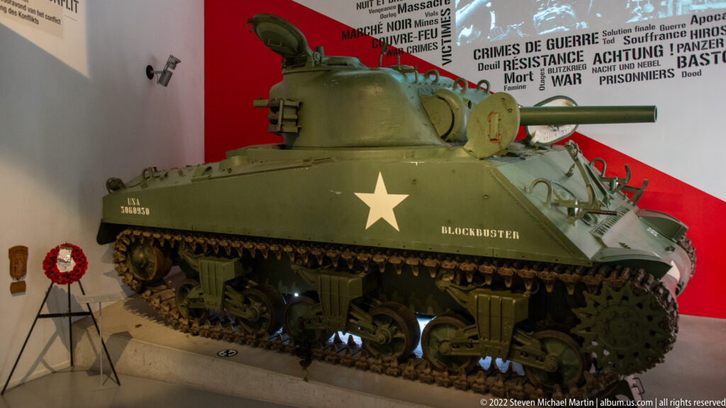 Olive-drab Sherman tank named Blockbuster with white star, turret, gun barrel, tracks, and suspension visible indoors.