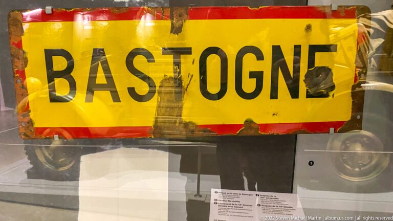 Weathered yellow road sign with red border and large black word 'BASTOGNE' in a museum case.
