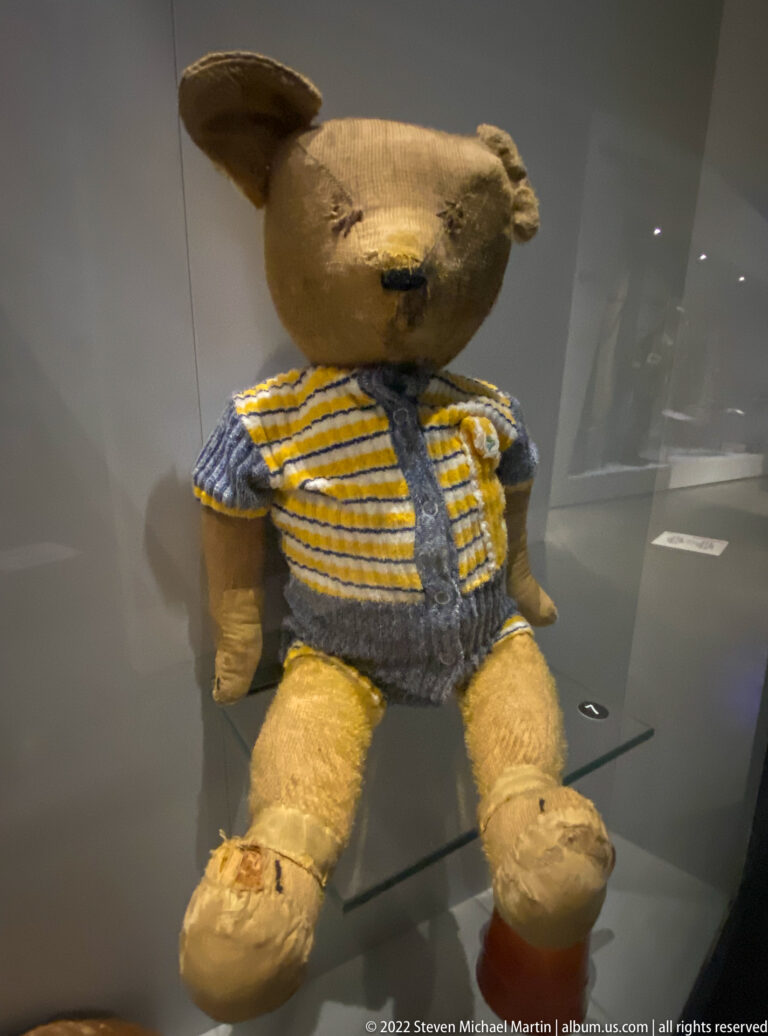 Worn tan teddy bear wearing a blue-and-yellow striped knitted outfit in a museum case.