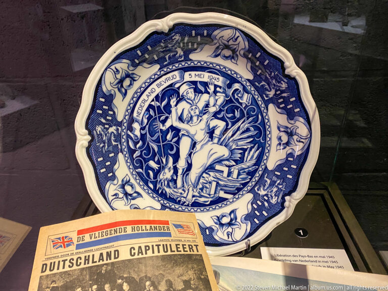 Blue-and-white commemorative plate dated 5 May 1945 above a Dutch newspaper announcing Germany's capitulation.