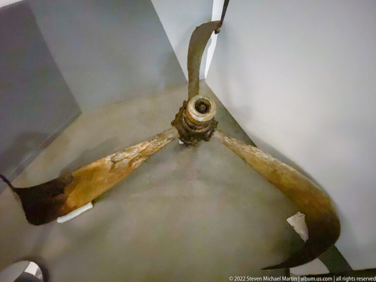 Corroded three-blade aircraft propeller with damaged hub mounted vertically in a display case.