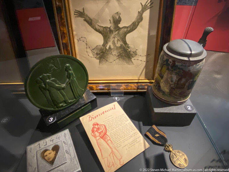 Display case with framed drawing, green relief plaque, ceramic stein, portrait, printed card, and medal.