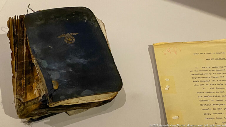 Severely damaged dark-covered book with warped pages beside a typed document in a museum display.