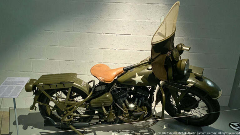 Olive-drab military motorcycle with tall windshield, leather seat, rear rack, and white star on the fuel tank.